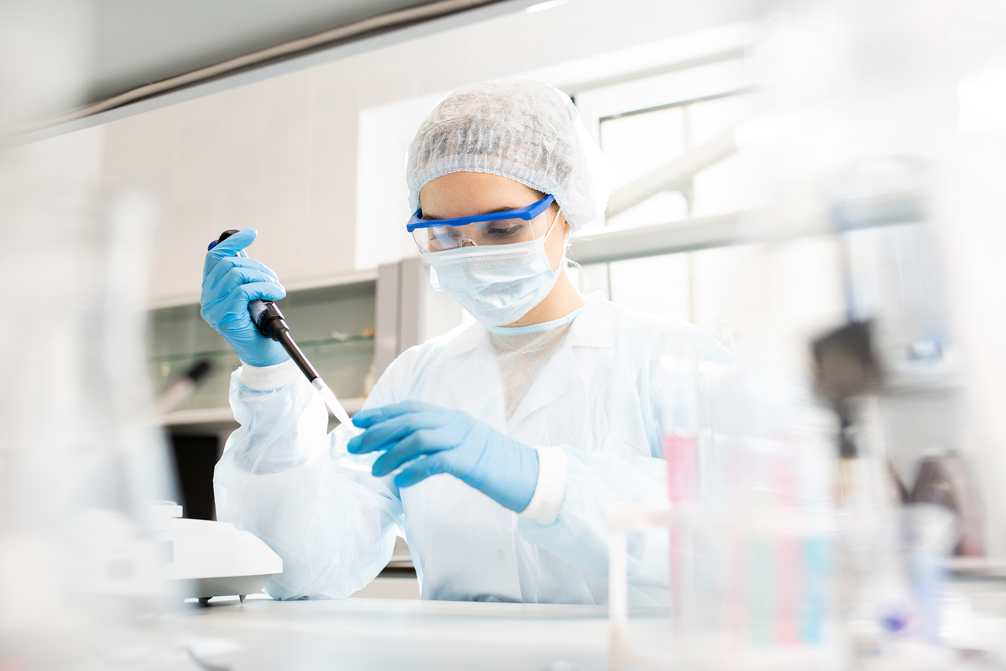 Serious concentrated female microbiologist in sterile clothing and safety goggles sitting at table and dropping reagent in petri dish while doing research in laboratory